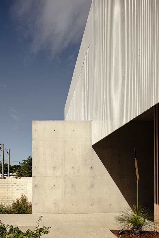 Contemporary architectural exterior featuring a tall white vertically ribbed facade, a smooth exposed concrete wall, and a deep dark recessed entrance or passage. The composition emphasizes sharp geometric forms, strong sunlight and shadow, and minimalist landscaping with sculptural grasses. A low pale masonry boundary wall and blue sky are visible in the background.