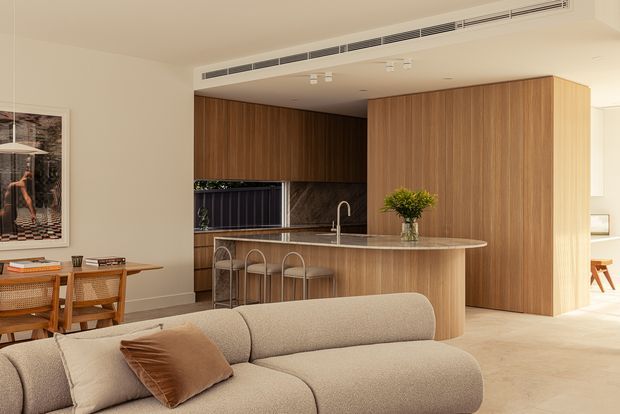 Contemporary open-plan living and kitchen interior with extensive warm timber cabinetry, a curved stone kitchen island with integrated sink and bar seating, and a neutral upholstered sofa in the foreground. A compact dining area and artwork sit to the left, while concealed or integrated storage creates a clean architectural backdrop. The space is softly lit and refined, with a warm neutral palette.