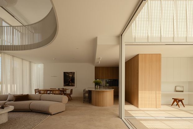 Warm, refined contemporary open-plan interior combining a living room, dining area and kitchen. The space features a large curved sectional sofa, dining table, rounded timber kitchen island, extensive pale cabinetry, a sculptural curved balustrade to an upper level, and large glazed openings with vertical blinds. Soft natural light creates patterned shadows across the pale stone-toned floor, while timber joinery adds warmth.