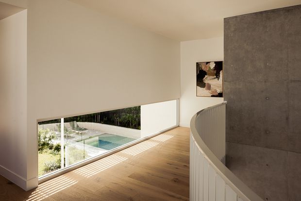 A minimalist upper-level hallway or landing with warm timber flooring, smooth off-white walls, and a curved white balustrade overlooking a lower level. Full-height glazing brings in views of a landscaped courtyard and pool, while a large exposed grey concrete wall forms a strong feature surface. Sunlight creates linear shadows across the floor.