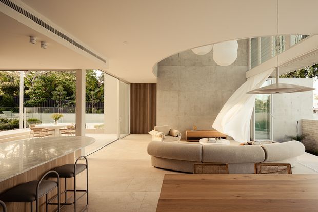 A bright, open-plan contemporary living, dining and kitchen interior with floor-to-ceiling glazing, a curved kitchen island, sculptural suspended rangehood, curved sectional sofa and warm timber dining table. A tall exposed concrete feature wall and flowing sheer curtain create a soft contrast with the minimal cream-toned finishes. The space opens toward a leafy outdoor setting.