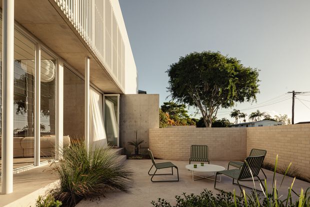 A contemporary residential courtyard or terrace viewed from outside, featuring a pale rendered or concrete two-storey facade with extensive floor-to-ceiling glazing, slim vertical columns, and a screened upper-level balcony. The paved outdoor area is enclosed by a light brick boundary wall and furnished with modern outdoor lounge chairs and a small round table, with clipped landscaping and ornamental grasses adding greenery.