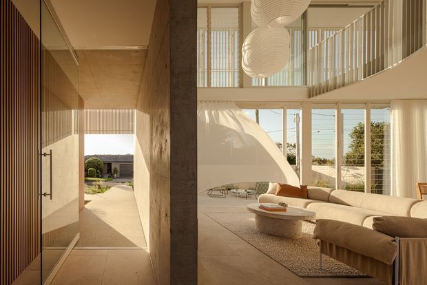 A refined open-plan interior combining a sculptural living room with a tall glazed void and an adjoining entry passage. Warm stone or concrete-toned walls and flooring, vertical timber screening, expansive floor-to-ceiling glazing, and a curved upper-level balustrade create a serene, light-filled contemporary space. The living area includes a large neutral sectional sofa, textured rug, low stone coffee table, and oversized woven pendant lights. A landscaped exterior is visible through the entry and glazing.