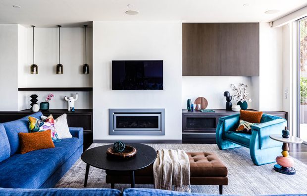 Bright contemporary living room with a large blue sectional sofa, teal armchair, black round coffee table, ottoman, wall-mounted television, built-in fireplace, dark timber cabinetry, and a large vertical timber wall panel. Floor-to-ceiling glazing on the right brings in natural light, while colourful cushions and sculptural accessories add vibrant accents.