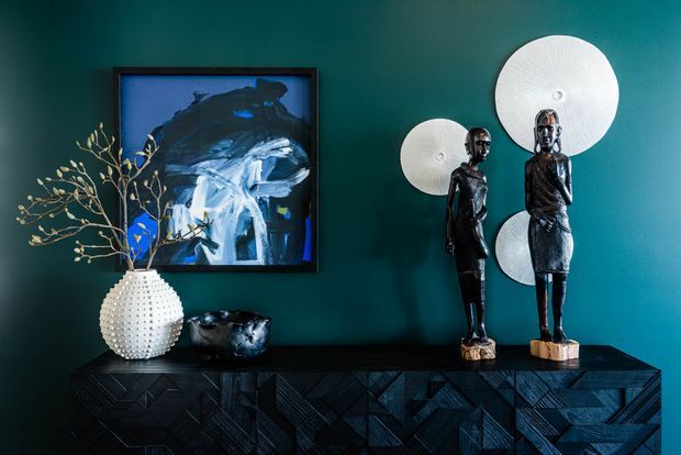 A moody, highly styled interior vignette featuring a dark teal painted wall, a black geometric-patterned sideboard, abstract artwork in a black frame, a white textured vase with branches, sculptural dark vessels, two dark figurines, and overlapping circular white wall sculptures.