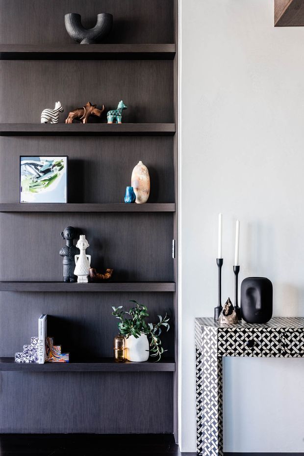 A refined interior vignette featuring a dark timber built-in shelving unit displaying sculptural objects, books, artwork, and plants. Beside it is a slim console table with a striking black-and-white geometric mosaic tiled surface and legs, styled with candles and decorative objects. The palette is high-contrast, combining dark wood, black accents, white walls, and patterned tile.