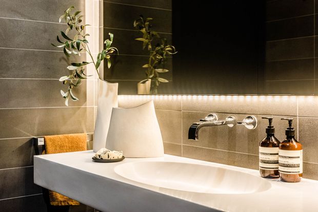 Contemporary bathroom vanity featuring a sculptural white integrated basin, wall-mounted chrome tapware, large dark mirror, warm under-cabinet lighting, and large-format grey wall tiles. Amber bathroom product bottles, a towel, and a potted branch add subtle warmth and greenery.