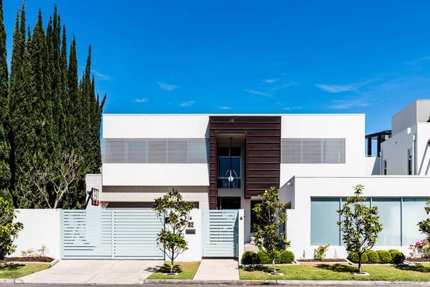 Street-facing view of a contemporary two-storey residential facade with crisp white rendered walls, broad horizontal glazing, a recessed central entry, dark timber-look vertical cladding, and a white garage volume. A low fence and landscaped front garden frame the property under a clear blue sky.