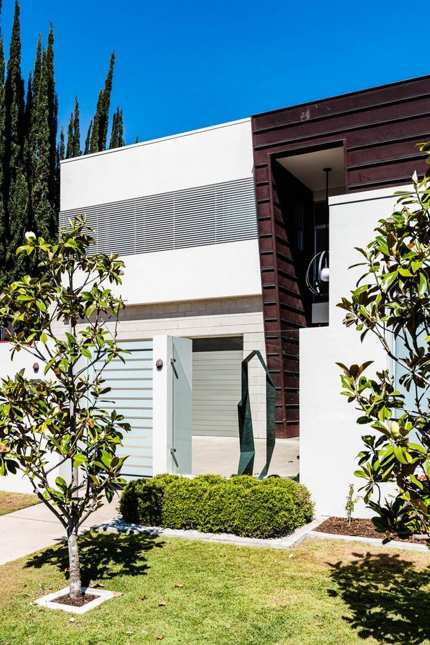 Bright exterior view of a contemporary flat-roofed building with crisp white rendered walls, horizontal louvre detailing, pale masonry at the entry, and a dark timber-toned cladding frame around a recessed doorway. The landscaped frontage includes clipped shrubs, small ornamental trees, lawn, and a paved path under a clear blue sky.