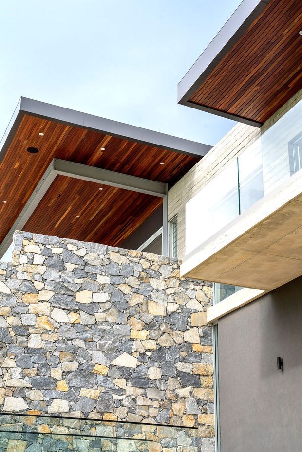 Upward-looking architectural exterior view of a contemporary multi-level residence. The image highlights projecting concrete forms, warm timber-lined soffits with recessed downlights, large glazing, a transparent glass balustrade, and a prominent irregular natural-stone masonry wall.