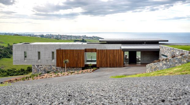A low-slung contemporary coastal residence set into a sloping site, viewed from above across a gravel foreground. The house combines exposed grey concrete masonry, warm vertical timber cladding, dark horizontal roof planes, extensive glazing, and rough stone retaining walls. Rolling green landscape and the ocean are visible beyond, under an overcast sky.