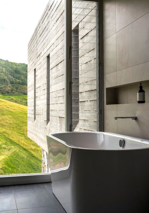 A contemporary bathroom with a freestanding sculptural bathtub positioned beside expansive floor-to-ceiling glazing. The room features large-format warm grey wall panels, dark grey floor tiles, a minimalist wall-mounted tap, and a recessed shelf with a black dispenser. Through the glazing, a textured concrete exterior wall and steep green hillside are visible. The composition emphasizes the strong connection between the refined interior and the natural landscape.