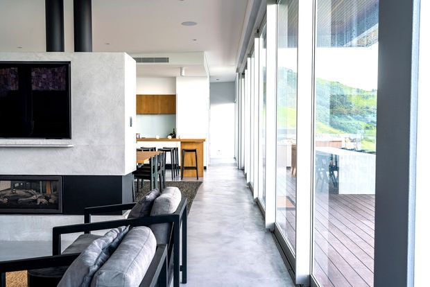 Bright contemporary open-plan living area with a large concrete fireplace and television feature wall, black-framed leather seating, and a kitchen with timber cabinetry and island bench beyond. Full-height glazing runs along the right side, opening visually to a timber deck and green landscape. The space has polished concrete flooring, a clean linear layout, and abundant natural light.