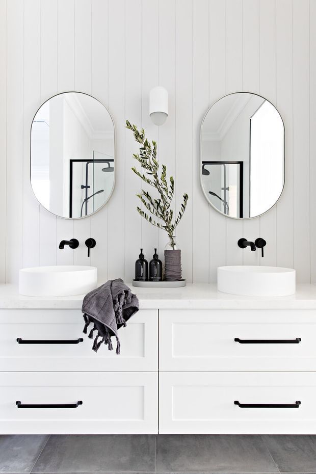 Bright, symmetrical contemporary bathroom vanity with twin white vessel basins, black wall-mounted tapware, arched round-edged mirrors, white vertical wall panelling, and a wide white built-in vanity with black bar handles. Grey large-format floor tiles and restrained styling create a clean, refined appearance.
