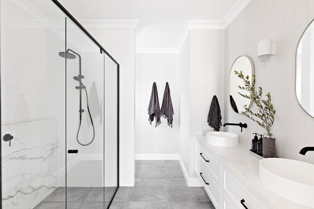 Bright contemporary bathroom with a walk-in glass shower on the left, large white vanity with vessel basins on the right, grey stone-look floor tiles, white vertical wall panelling, black fixtures, and a marble-look shower wall. Dark towels and a potted branch add contrast.