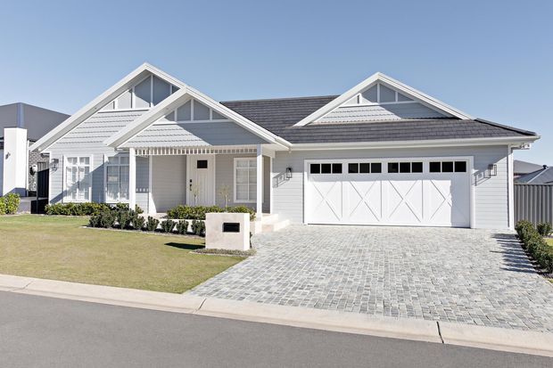 A bright suburban single-storey house exterior with a white weatherboard facade, multiple front-facing gables, grey tiled roofing, decorative gable ventilation details, a covered entry porch, and a large double garage. The front yard is neatly landscaped with lawn and low shrubs, while the driveway is finished in light grey pavers.