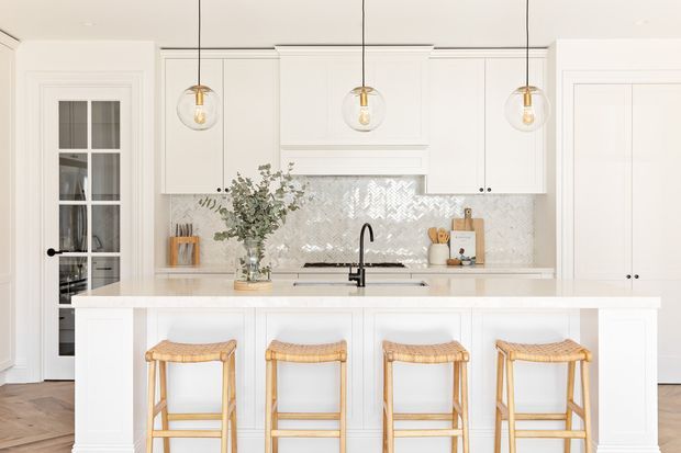 Bright, refined white kitchen with a large island and four woven-seat timber stools. The room features white shaker-style cabinetry, a pale patterned tile splashback, light stone benchtops, a black gooseneck tap, brass-and-glass pendant lights, and timber flooring. Glazed double doors and tall pantry cabinetry are visible at the sides.