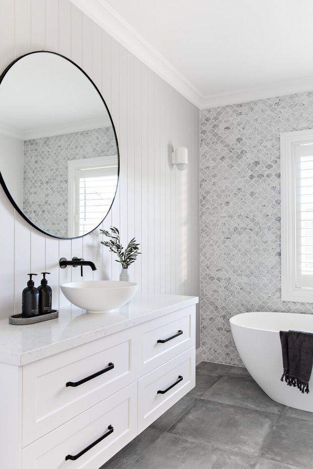 Bright, refined bathroom featuring a white floating-style vanity with black hardware, white vessel basin and black tapware, a large round black-framed mirror, vertical white wall panelling, grey scallop-pattern wall tiles, grey tiled flooring, and a freestanding white bathtub beside a shuttered window.