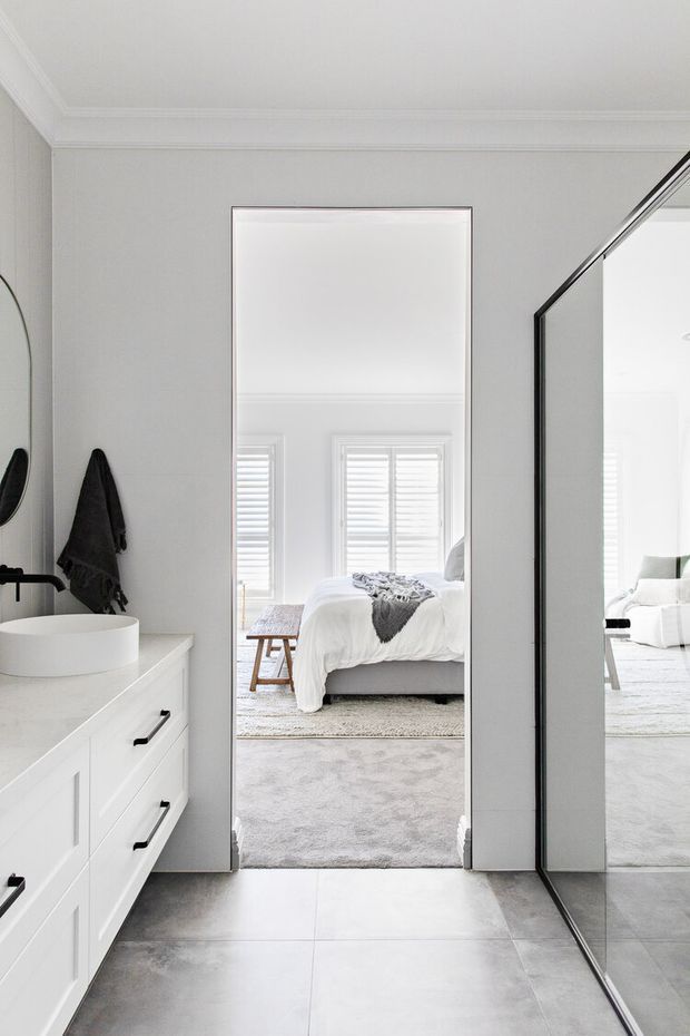 Bright, refined bathroom interior photographed toward an open doorway into a bedroom. The bathroom features a white floating vanity with black handles, a white vessel basin, large grey floor tiles, pale walls, and full-height mirrored wardrobe doors. The bedroom beyond has white bedding, plantation shutters, and a soft grey rug.