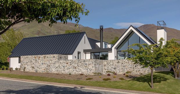 Exterior view of a contemporary gabled home set against rugged hills. The building combines dark standing-seam metal pitched roofs, crisp white rendered walls, large triangular gable glazing, and a low natural-stone boundary wall. Landscaped lawns and mature trees frame the street-facing facade.