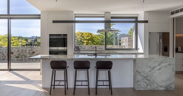 A bright, contemporary open-plan kitchen centered on a large island with three dark bar stools. The island features a pale marble-look waterfall end panel and a light stone benchtop, with vertically ribbed white cabinetry below. Full-height glazing and sliding doors open to a stone terrace, while integrated appliances and minimalist dark pendant lighting contribute to the clean, refined design. Greenery and distant hills are visible outside.