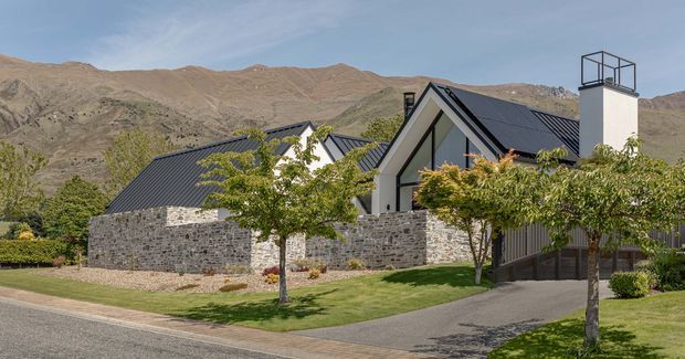 Exterior view of a contemporary mountain house with steep gabled roof forms, dark metal roofing, pale rendered gable walls, and extensive grey natural-stone walls. A tall chimney with a metal cap rises above the roofline. The property has a landscaped front lawn, young deciduous trees, paved driveway and walkway, and dramatic dry mountain terrain in the background.