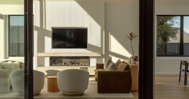A warm, contemporary living room viewed through a large glazed opening, featuring a linear fireplace and wall-mounted television set within a full-height vertically ribbed feature wall. Curved upholstered lounge chairs, a brown sofa, a low timber coffee table, and a textured rug create a refined seating area. Strong afternoon sunlight casts geometric shadows across the room. A dining area and exterior roofline are visible beyond the glazing.