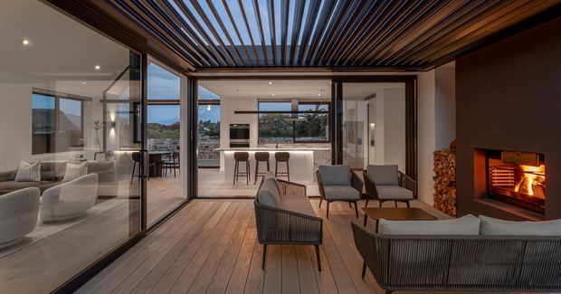 A contemporary indoor-outdoor living space with full-height glazed sliding doors, a timber-lined floor and ceiling, and a dramatic slatted pergola overhead. The room includes lounge seating, a dining or bar area visible beyond the glazing, and a built-in open fireplace with stacked firewood. The warm timber, dark framing, and evening light create a refined, inviting atmosphere.