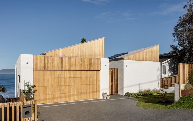 Exterior view of a contemporary residential property with white rendered walls, prominent vertical timber-clad garage doors and upper wall planes, sharply angled mono-pitched roof forms, timber privacy screens and fencing, and a paved driveway. A body of water is visible beyond the site, with landscaped lawn and planting in the foreground.