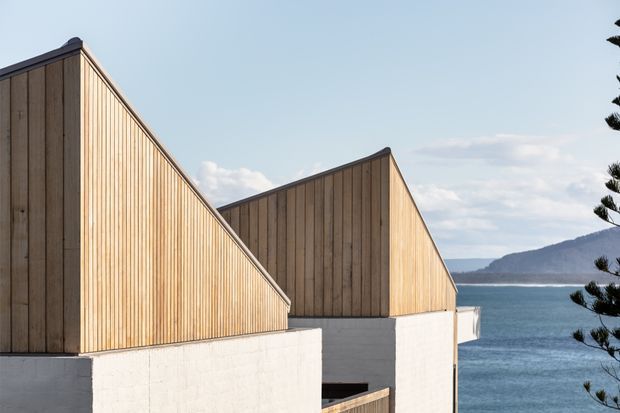 Exterior architectural view of a modern coastal building composed of angular, gabled volumes clad in vertical timber boards above white masonry walls. The structure overlooks a blue ocean with a mountainous shoreline in the background.