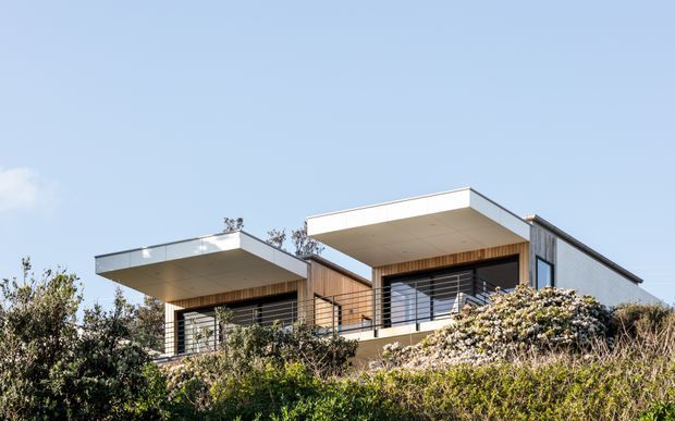 Exterior view of a contemporary two-storey residence set into a landscaped, sloping site. The house features crisp flat roof planes with deep cantilevered overhangs, light rendered or concrete walls, warm vertical timber cladding around the upper-level openings, dark metal-framed glazing and horizontal steel balustrades. Dense coastal-style planting partially screens the lower level.