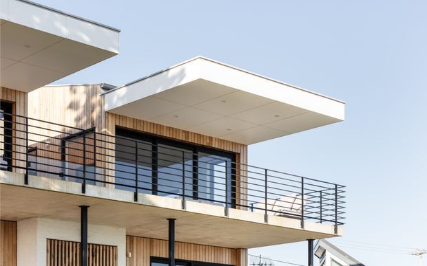 Contemporary multi-level residential facade with projecting flat roof slabs, warm vertical timber cladding, large glazed openings, and black metal balcony balustrades. The image focuses on the building exterior in bright daylight.