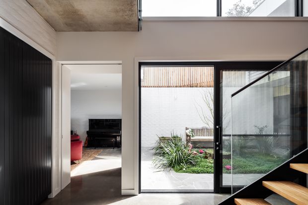 A bright contemporary interior overlooking a small landscaped courtyard. The space features a double-height glazed opening with black-framed sliding doors, a black open-riser staircase with timber treads, dark flooring, white walls, and a concrete soffit. A piano and red seating are visible in the adjoining room, while a high-level window brings in additional natural light.