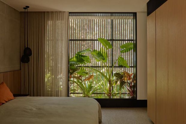 Warm, softly lit bedroom with a low-profile bed, textured neutral bedding, full-height curtains, and a large black-framed window overlooking lush tropical planting. Timber wall panelling and built-in cabinetry create a calm, natural interior, with a dark pendant light and a small orange accent cushion.
