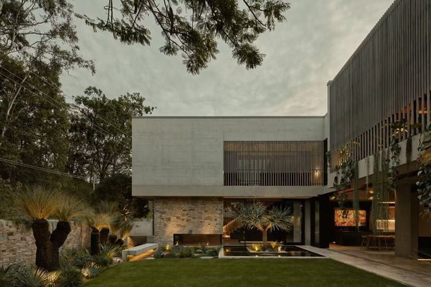 Contemporary multi-level residence set within a landscaped garden, viewed from the exterior courtyard. The composition features a rectilinear pale concrete upper volume, textured stone masonry at ground level, dark vertical screened cladding and extensive glazing. Warmly lit outdoor living areas open onto a lawn with sculptural planting and a shallow reflective water feature. Mature trees frame the scene under an overcast sky.