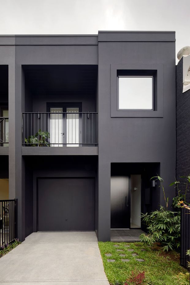 A narrow contemporary townhouse facade in dark charcoal grey, viewed from the front. The two-storey building has a flat roofline, minimalist rectangular forms, a recessed upper balcony with slim black metal balustrade, a projecting framed window, an integrated single garage, and a recessed entry. A concrete driveway and stepping stones through a small planted garden lead to the entrance.