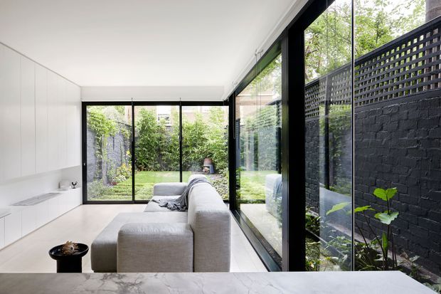 Bright contemporary open-plan living space with a minimalist white built-in kitchen along the left wall, a large pale sectional sofa, and expansive black-framed floor-to-ceiling glazing opening views to a secluded green courtyard. A dark painted brick boundary wall with perforated screening lines the side garden, while a pale stone island or benchtop occupies the foreground.