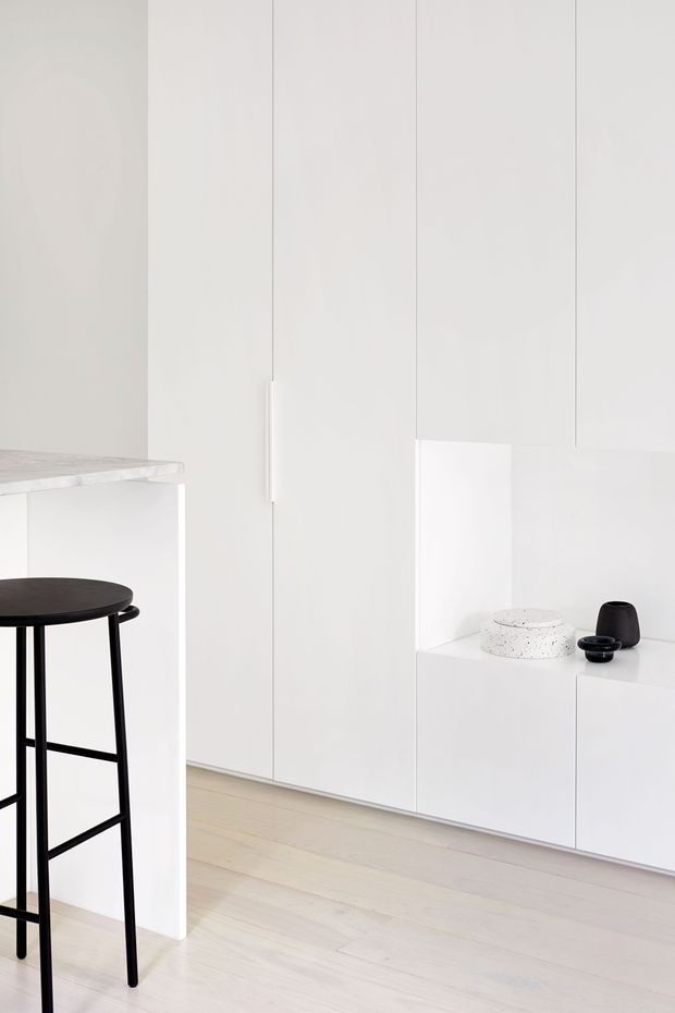 A minimalist white built-in interior with full-height flat-panel cabinetry, an open recessed display niche, and pale timber flooring. At left is a white stone or composite benchtop with a tall black stool. The space is bright, sparse, and monochromatic, with small dark decorative objects and a speckled light vessel in the niche.