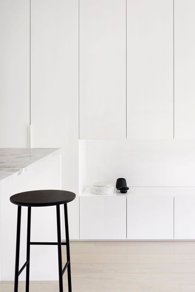 A minimalist white kitchen with full-height handleless cabinetry, a pale stone benchtop, and light timber flooring. A black bar stool sits at the island, while a recessed open shelf displays a few small objects. The composition is bright, clean, and highly restrained, with slim vertical cabinet seams and a monochromatic palette.