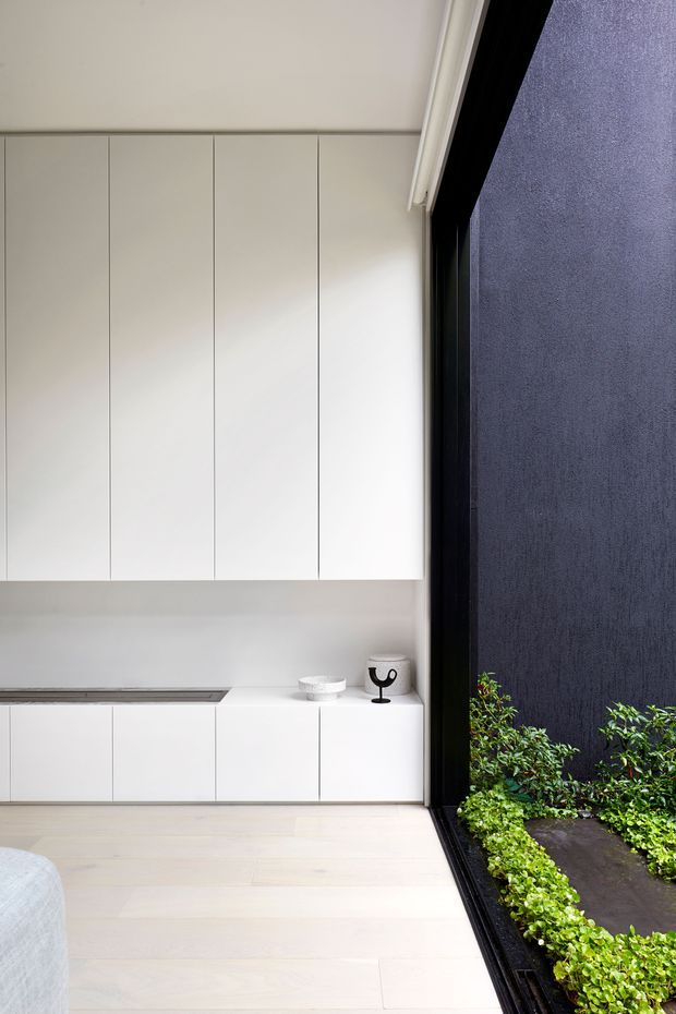 A minimalist white built-in joinery wall with tall handleless cabinets above low cabinetry, opening onto a narrow planted courtyard through a large black-framed glazed door or window. Pale timber flooring, a slim grey benchtop or ledge, and sparse black-and-white decorative objects create a clean, refined interior with strong indoor-outdoor connection.