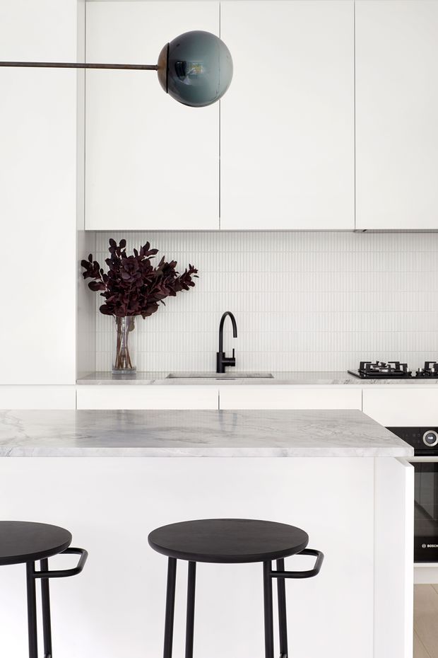 A bright, minimalist kitchen with white flat-panel cabinetry, a pale vertically ribbed tile splashback, dark tapware and appliances, and a large light stone island with black bar stools. A spherical wall light and burgundy foliage add restrained accents.