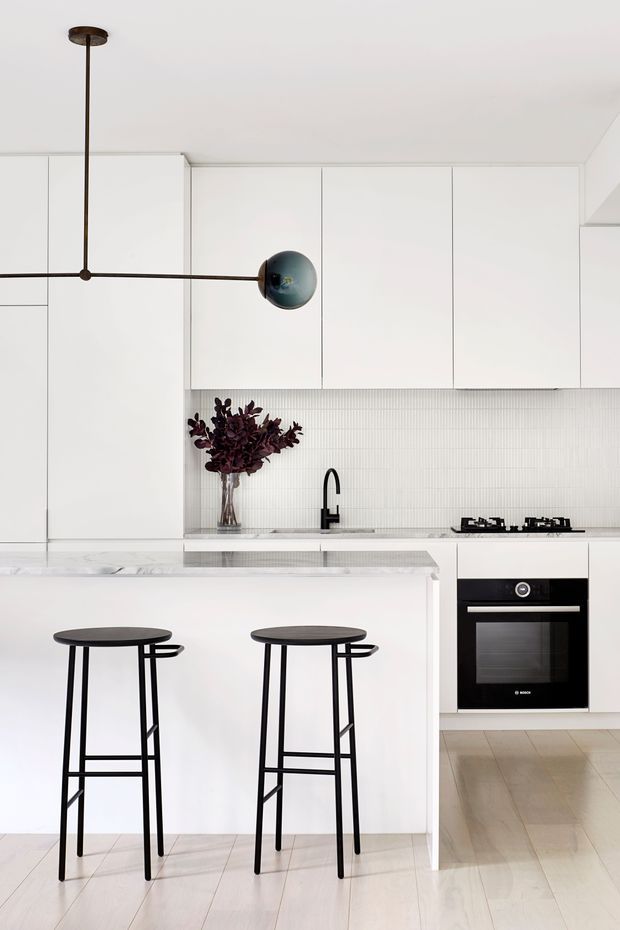A bright, minimalist contemporary kitchen with extensive white flat-panel cabinetry, a white island with a pale stone benchtop, integrated black gas cooktop and built-in oven, and a narrow white vertical-tile splashback. Two slim black bar stools sit at the island, complemented by a sculptural dark pendant light. Pale timber flooring and restrained styling create a clean, high-contrast interior.