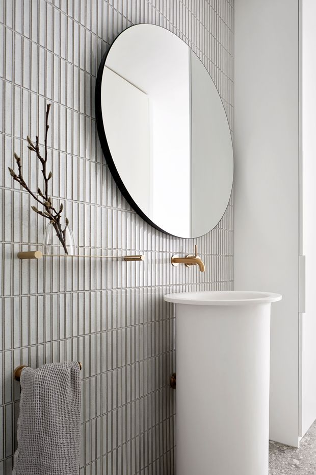 A refined contemporary bathroom featuring a wall-mounted oval mirror with a dark edge, vertically ribbed light-grey tiled walls, and brushed brass wall-mounted tapware and towel rail. A freestanding white basin sits in front of a white wall, with a textured grey hand towel and a subtle terrazzo-like floor visible at the bottom right. The space is bright, minimal, and carefully detailed.