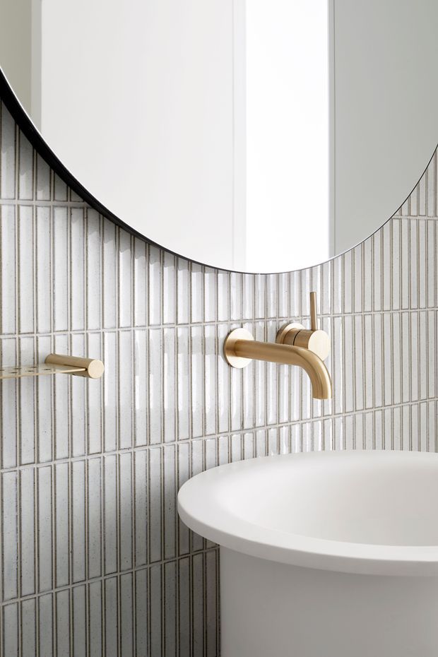 Close-up of a refined bathroom vanity area featuring vertically aligned glossy light-grey and white rectangular wall tiles, a curved-edge mirror, a white round vessel basin, and brushed brass wall-mounted tapware. The composition is clean and contemporary with soft neutral tones and warm metallic accents.