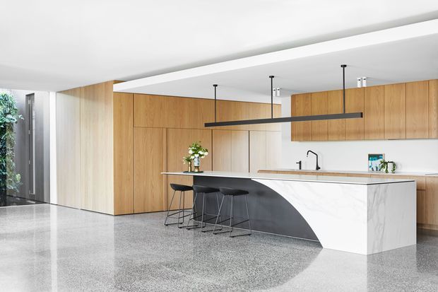 A bright, spacious contemporary kitchen with extensive warm timber cabinetry, a large white marble-look island with a dramatic curved dark-grey side panel, black bar stools, and a long linear pendant light. The space features polished grey terrazzo-style flooring, white walls and ceiling, integrated appliances concealed behind cabinetry, and a large opening toward greenery at the left.