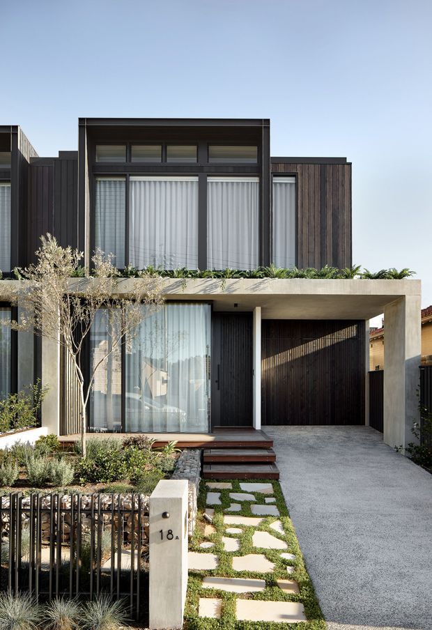 Front exterior view of a narrow contemporary multi-storey townhouse, featuring dark vertical timber-look cladding, large glazed openings with sheer curtains, a concrete-framed covered entry and integrated carport, landscaped front garden, and a stepping-stone path.