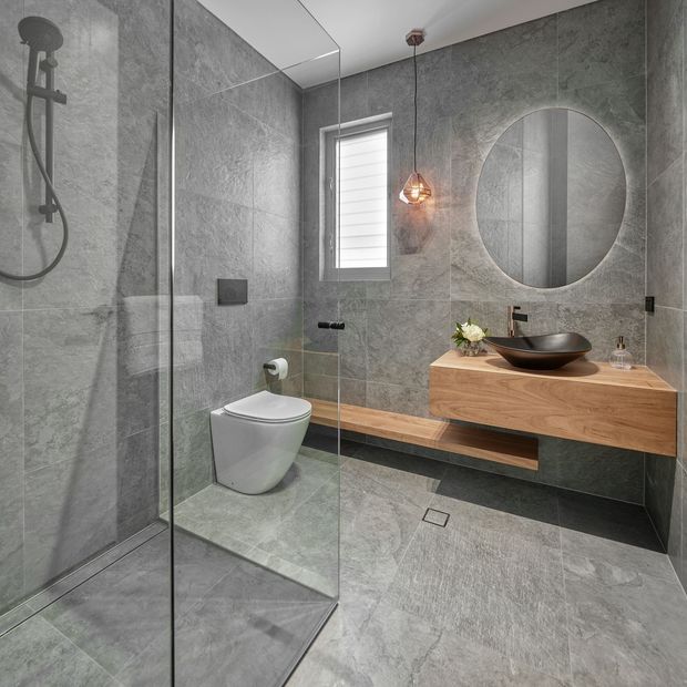Contemporary bathroom with full-height grey stone-look wall and floor tiles, a frameless glass shower enclosure, wall-hung toilet, floating timber vanity with a black vessel basin, round mirror, black tapware, and a warm pendant light. The palette is grey, timber, black, and warm white.