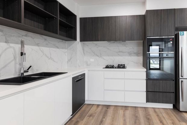 A modern L-shaped kitchen with white lower cabinetry, dark wood-grain overhead and tall cabinetry, a white marble-look splashback, black sink and tapware, integrated appliances, gas cooktop, and warm timber-look flooring.
