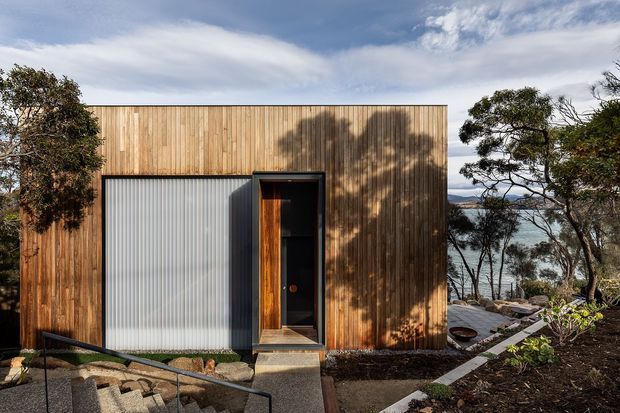 A compact contemporary flat-roofed building set into a landscaped, sloping site. The facade combines vertical natural timber boards with a translucent corrugated panel and a dark recessed entry door. Concrete stepping paths, boulders, native-looking vegetation, and glimpses of water beyond the trees create a secluded coastal setting.