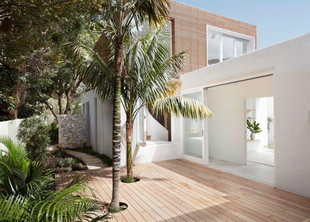 Bright contemporary house exterior with white rendered walls, extensive glass doors and windows, and a horizontal timber-lined upper facade. A broad timber deck leads through an open corner to the interior, while tropical palms and dense landscaping create a lush courtyard-like setting.